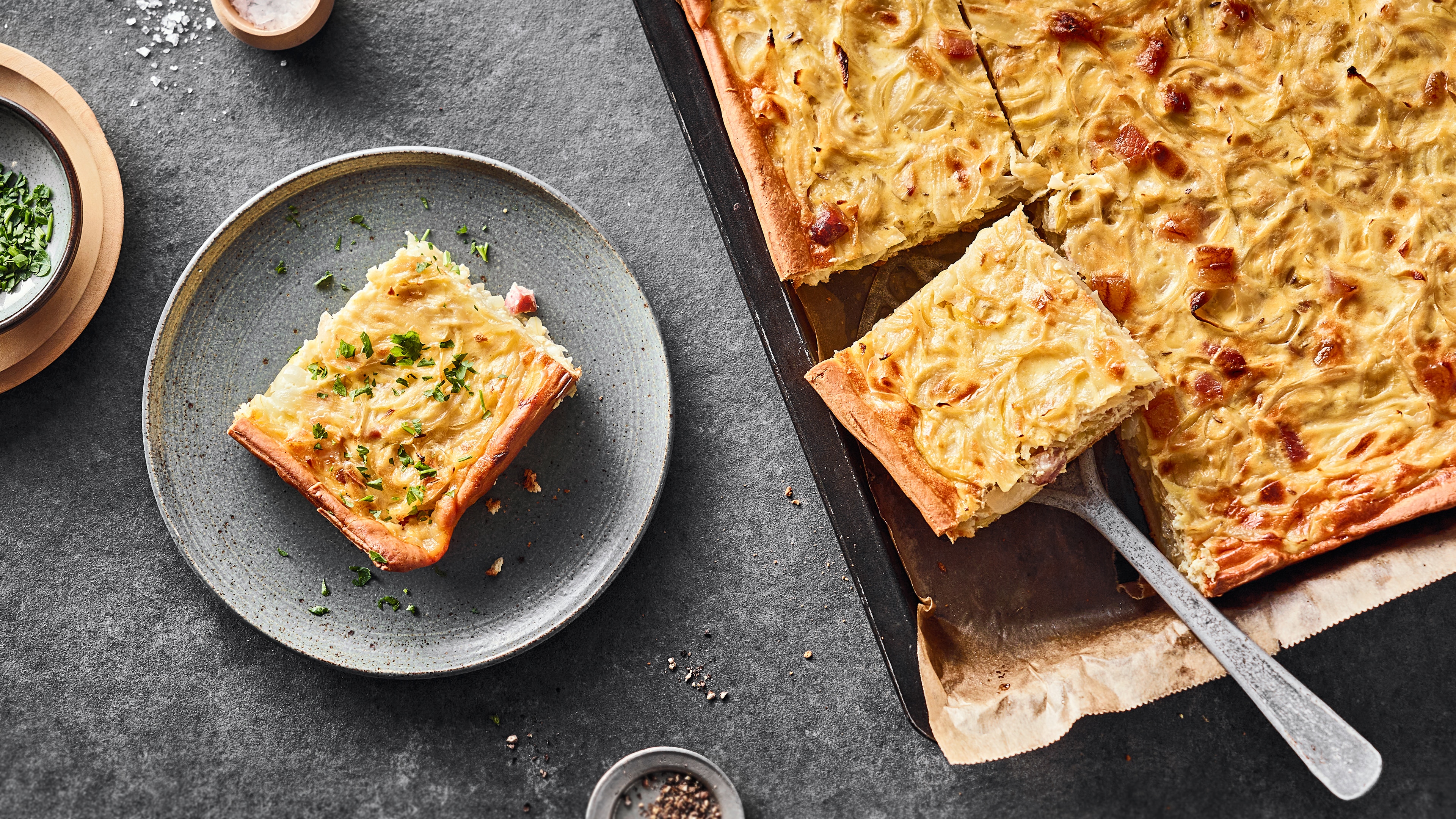Rezept für Zwiebelkuchen vom Blech Ein Teller mit einem Stück gelbgold gebackenen Zwiebelkuchen, getoppt mit frischer Petersilie