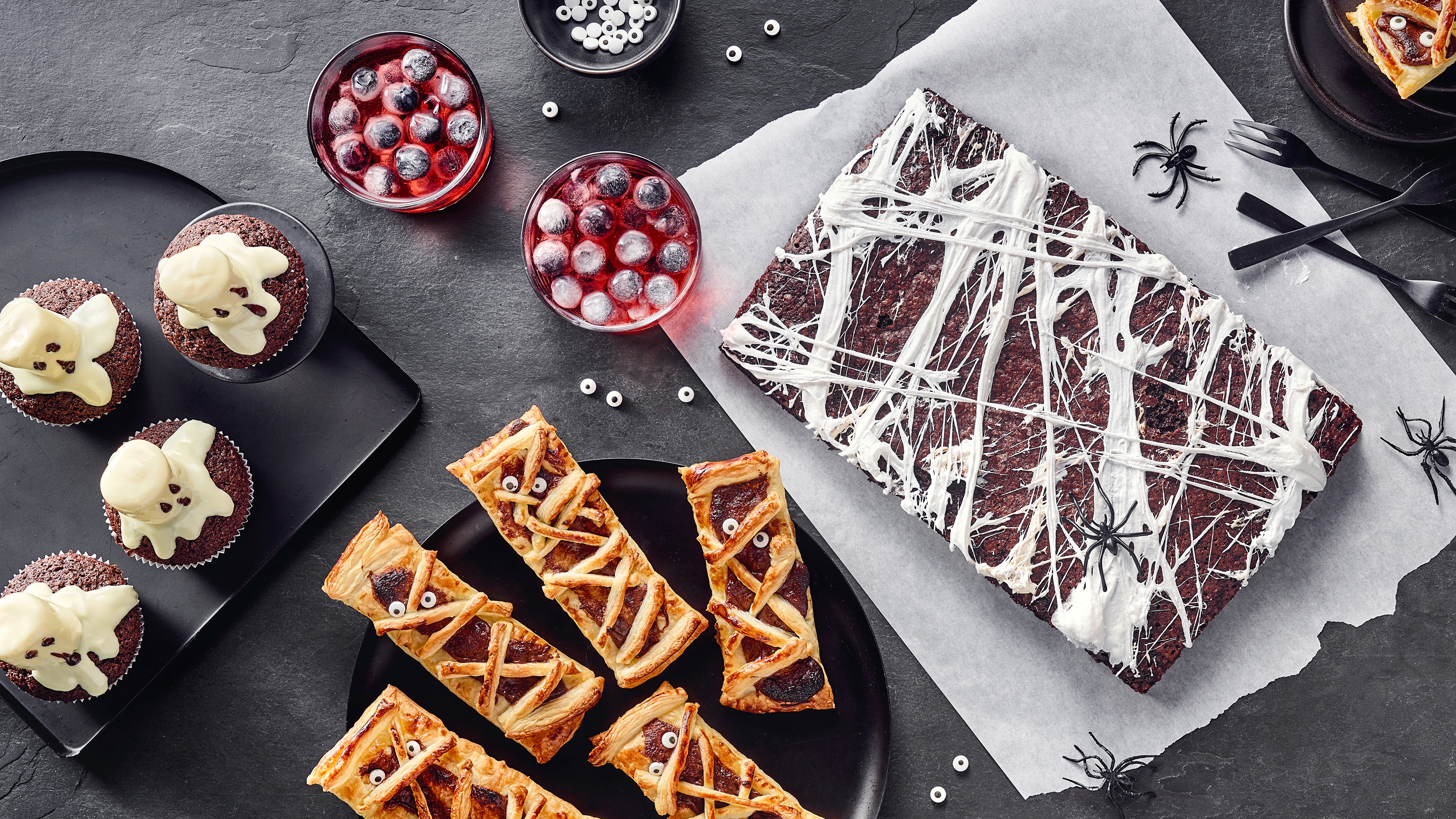 Ein großer Brownie mit weißer Verzierung, daneben Drinks mit Eiswürfeln sowie Muffins und Blätterteig-Gebäck