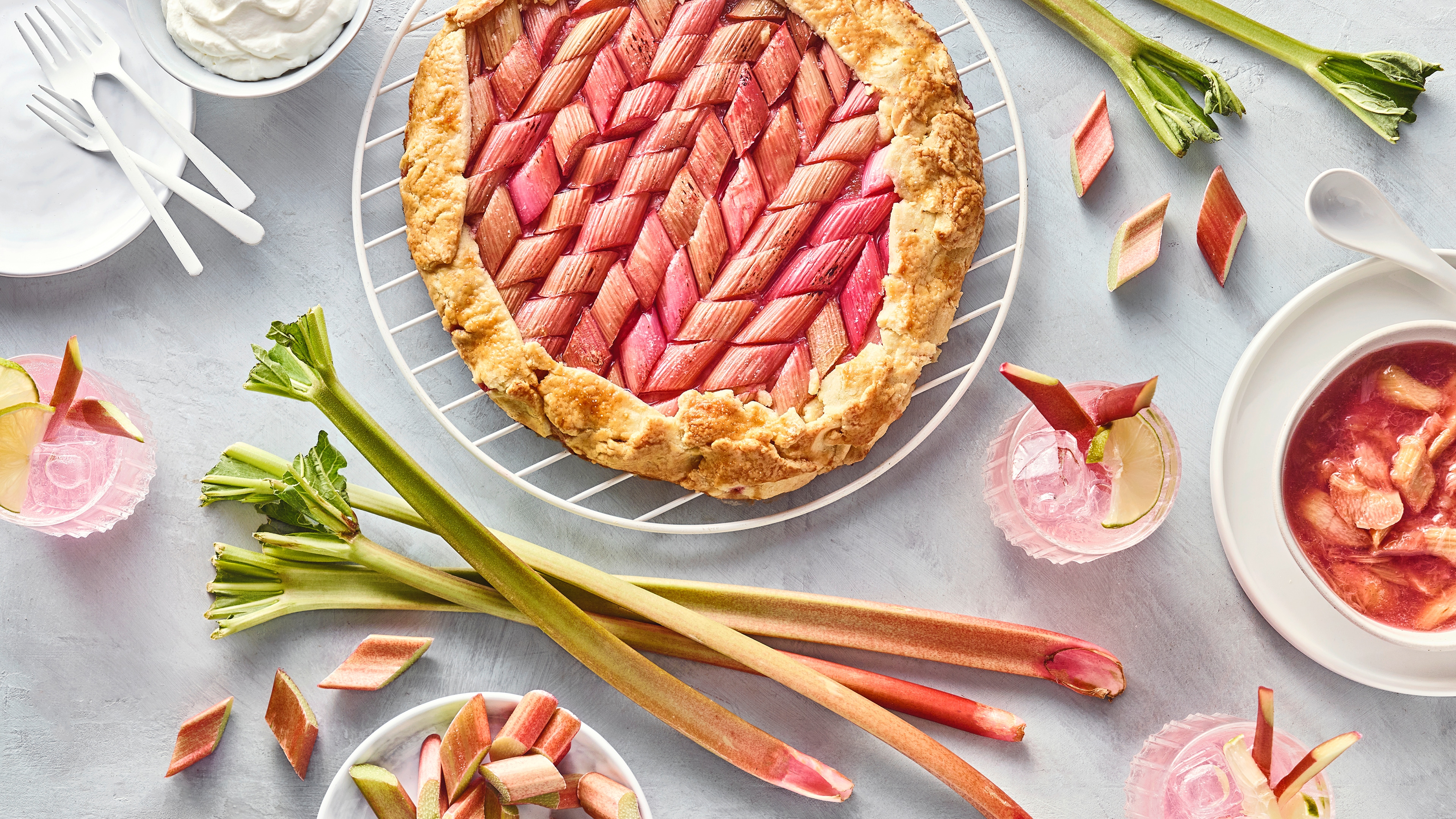 Großes Gebäck mit Rhabarberstücken, daneben Rhabarberstange, Gläser mit hellrosa Flüssigkeit und eine Schale mit Kompott