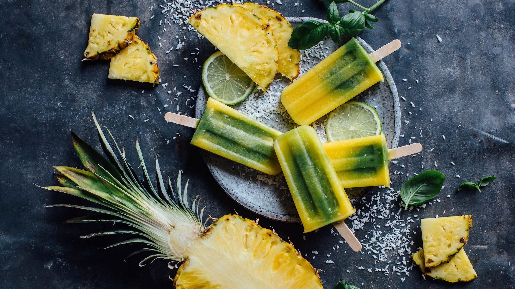 Vier gelb‑grüne Popsicles auf einem Teller, umgeben von aufgeschnittener Ananas; auf dem Teller liegen zusätzlich frische Limonen- und Ananasscheiben.