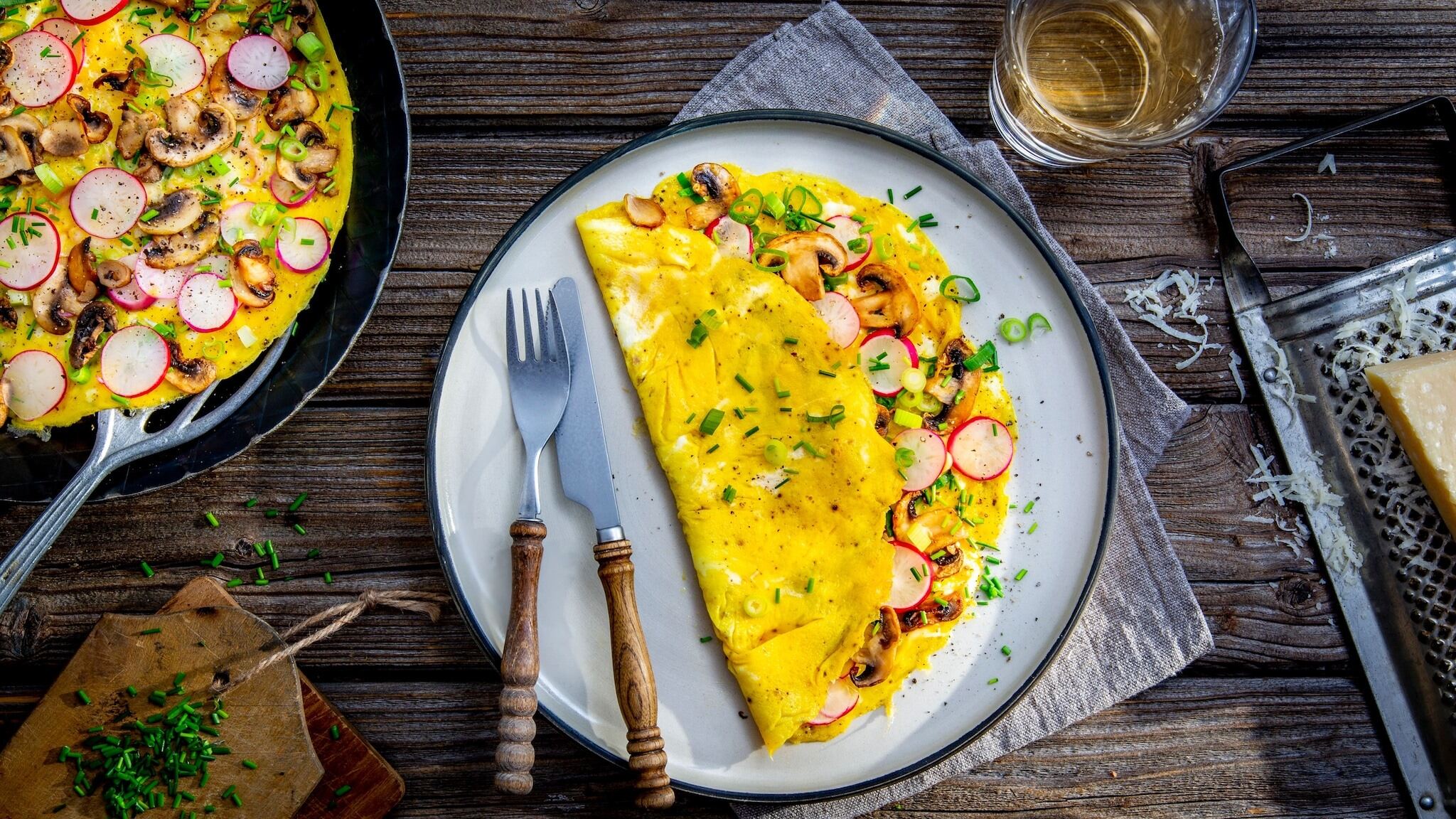 Eier-Omelette mit Radieschen und Schnittlauch. Links oben steht eine Pfanne mit einem weiteren Omelette. Rechts unten liegt eine Reibe mit Parmesan.