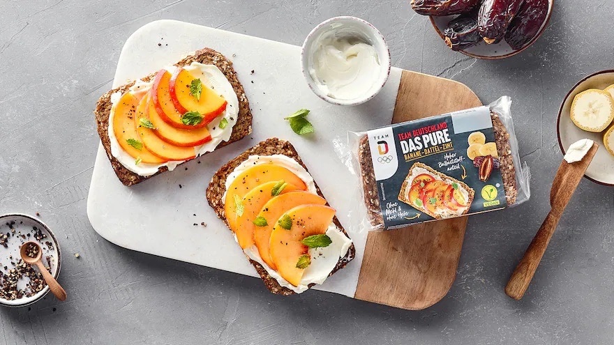 Zwei belegte Scheiben Schwarzbrot auf einem Brett. Außerdem eine geschlossene Packung Brot sowie Schälchen mit Datteln und Banane.