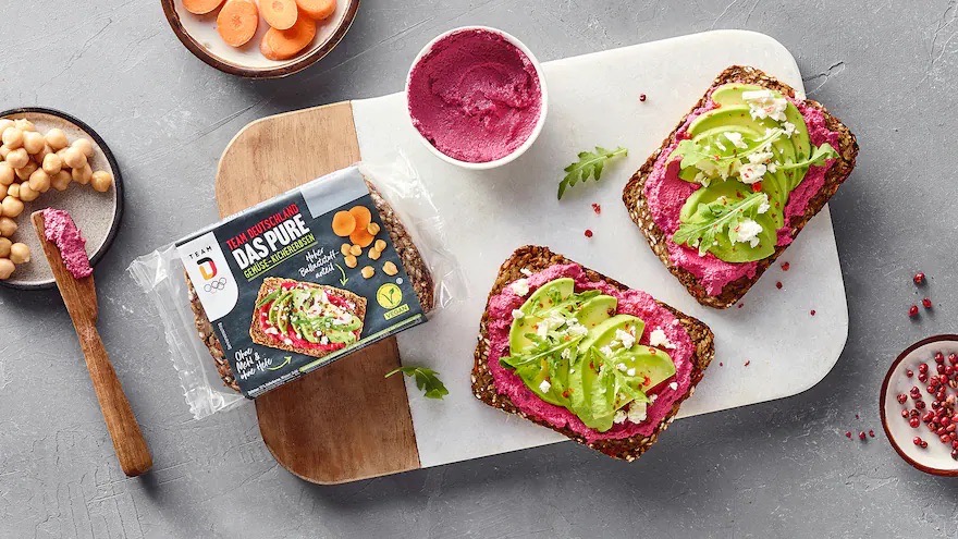 Zwei belegte Scheiben Schwarzbrot auf einem Brett. Außerdem eine geschlossene Packung Brot sowie Schälchen mit Mohrrüben, Kicherebsen und einer roten Creme.