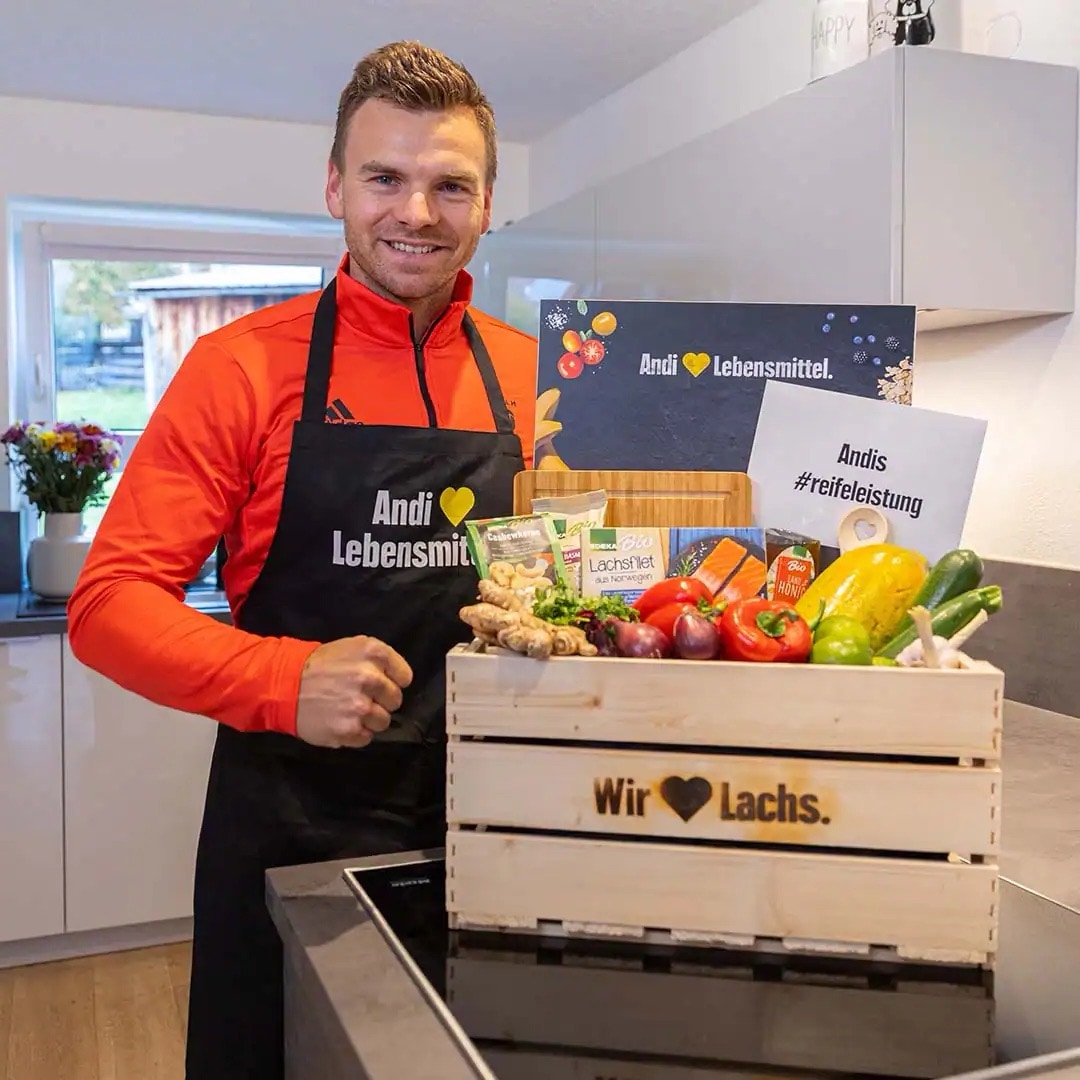 Junger Mann in Sportoberteil und EDEKA-Schürze. Auf der Herdplatte vor ihm eine Holzkiste mit Lebensmitteln. Auf einem Blatt Papier steht Andis Reifeleistung.