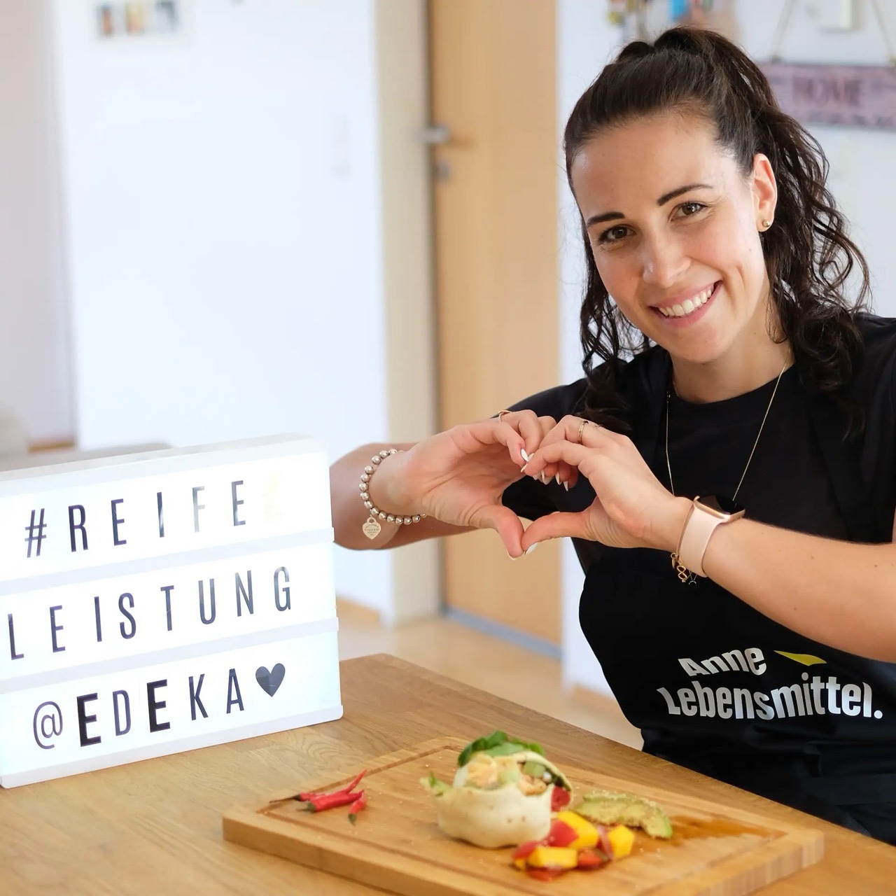 Junge Frau formt mit den Händen ein Herz. Vor ihr ein Schneidebrett mit Gemüse und ein Leuchtkasten mit dem Text: #reife Leistung @ EDEKA.