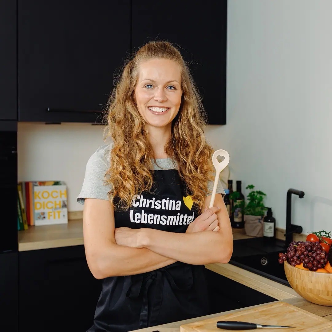 Junge Frau mit langen braunen Haaren steht lachen in einer Küche. Sie trägt eine schwarze Schürze mit Text: Christina liebt Lebensmittel.