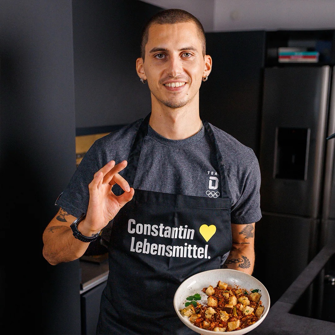 Junger Mann mit sehr kurzen Haaren und schwarzer Schürze mit Text: Constantin liebt Lebensmittel. Er hält einen tiefen Teller mit verarbeiteten Lebensmitteln.