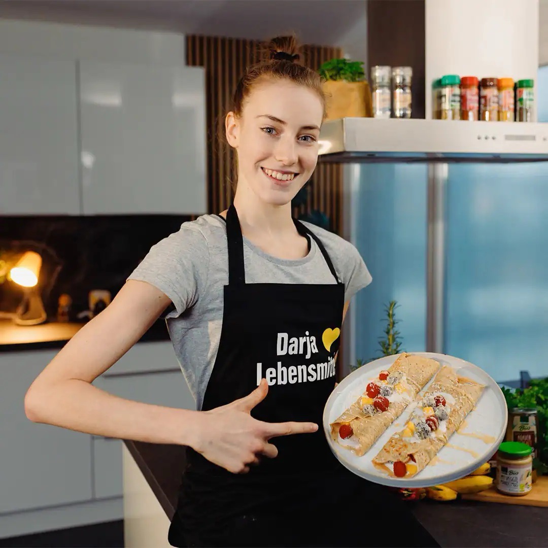 Junge Frau mit schwarzer EDEKA-Schürze präsentiert einen Teller mit eingerollten Pfannkuchen. Auf ihrer Schürze steht „Darja liebt Lebensmittel.“