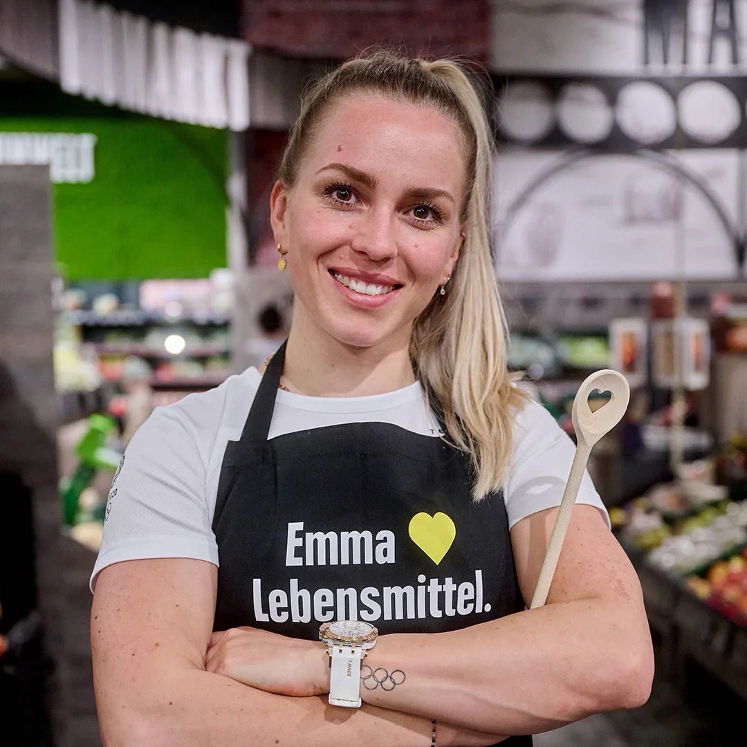 Junge Frau mit schwarzer Schürze, auf der „Emma liebt Lebensmittel“ steht. Sie hat die Arme vor sich verschränkt, auf ihrem Unterarm sind fünf Ringe tätowiert – das Symbol der Olympischen Spiele.