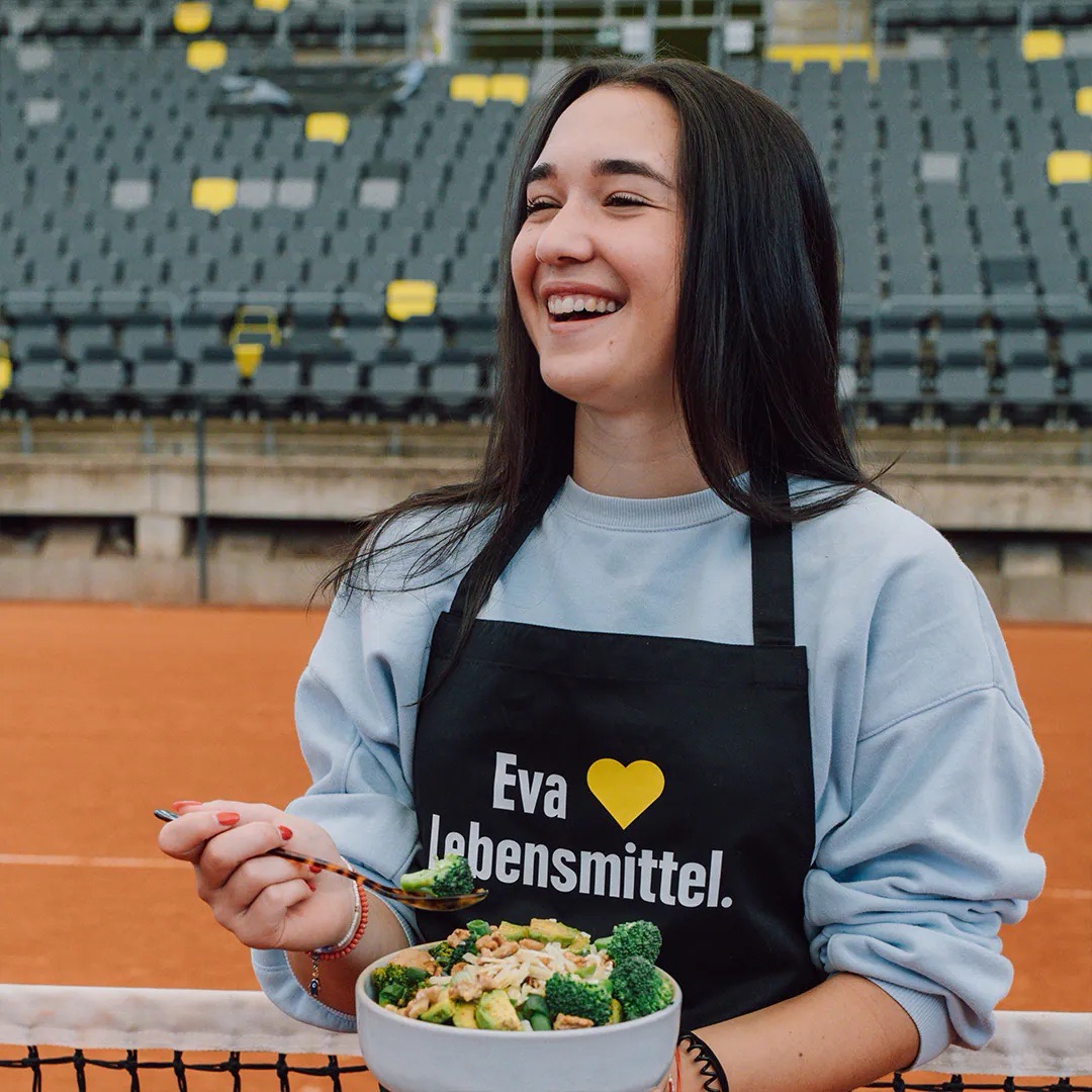 Junge Frau mit schwarzer EDEKA-Schürze auf der „Eva liebt Lebensmittel“ steht. Sie befindet sich am Netz auf einem Tennisplatz und hält eine Bowl mit Broccoli in der Hand.
