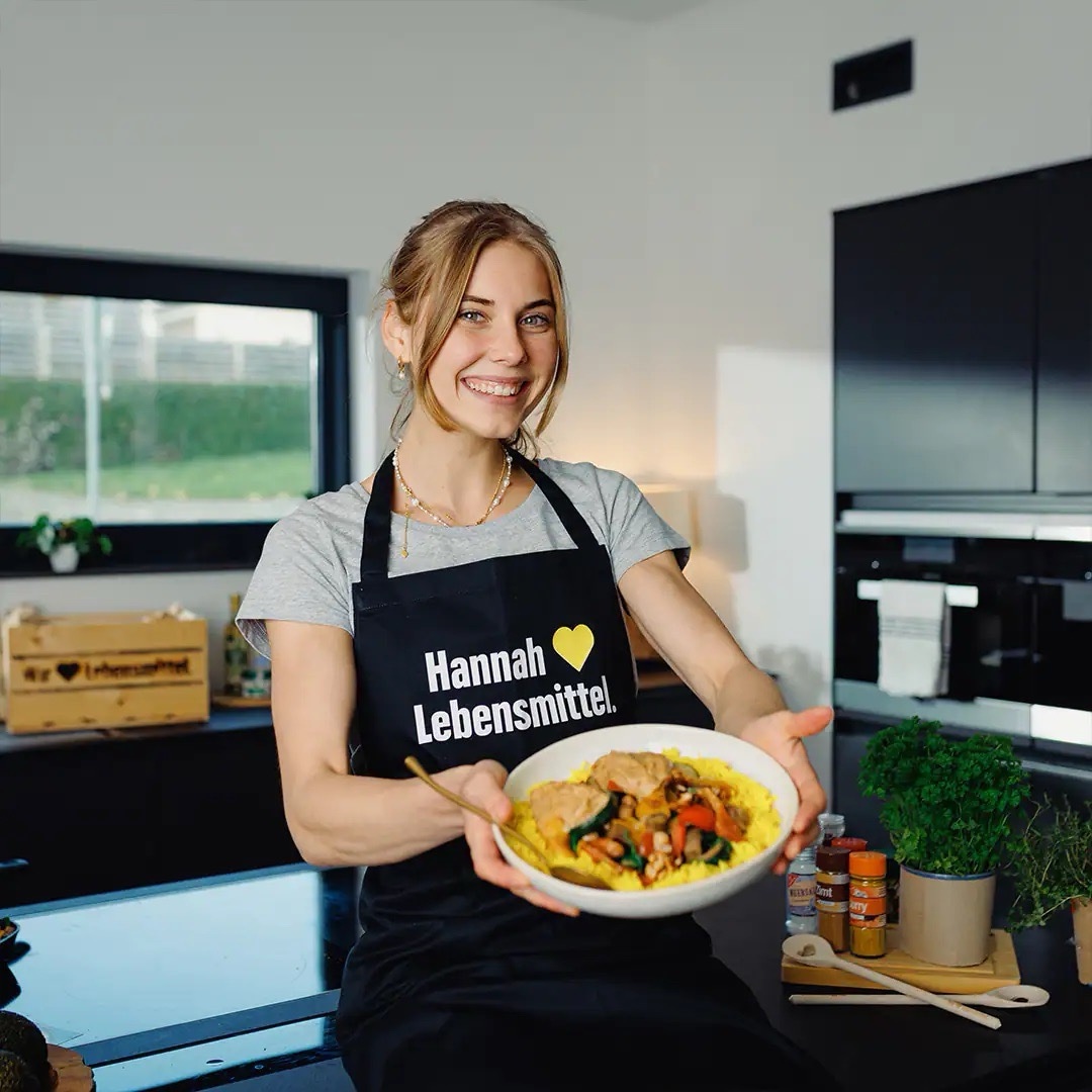 Junge Frau mit schwarzer EDEKA-Schürze, auf der „Hannah liebt Lebensmittel“ steht. Sie steht in einer modernen Küche und hält einen vollen, tiefen Teller vor sich.