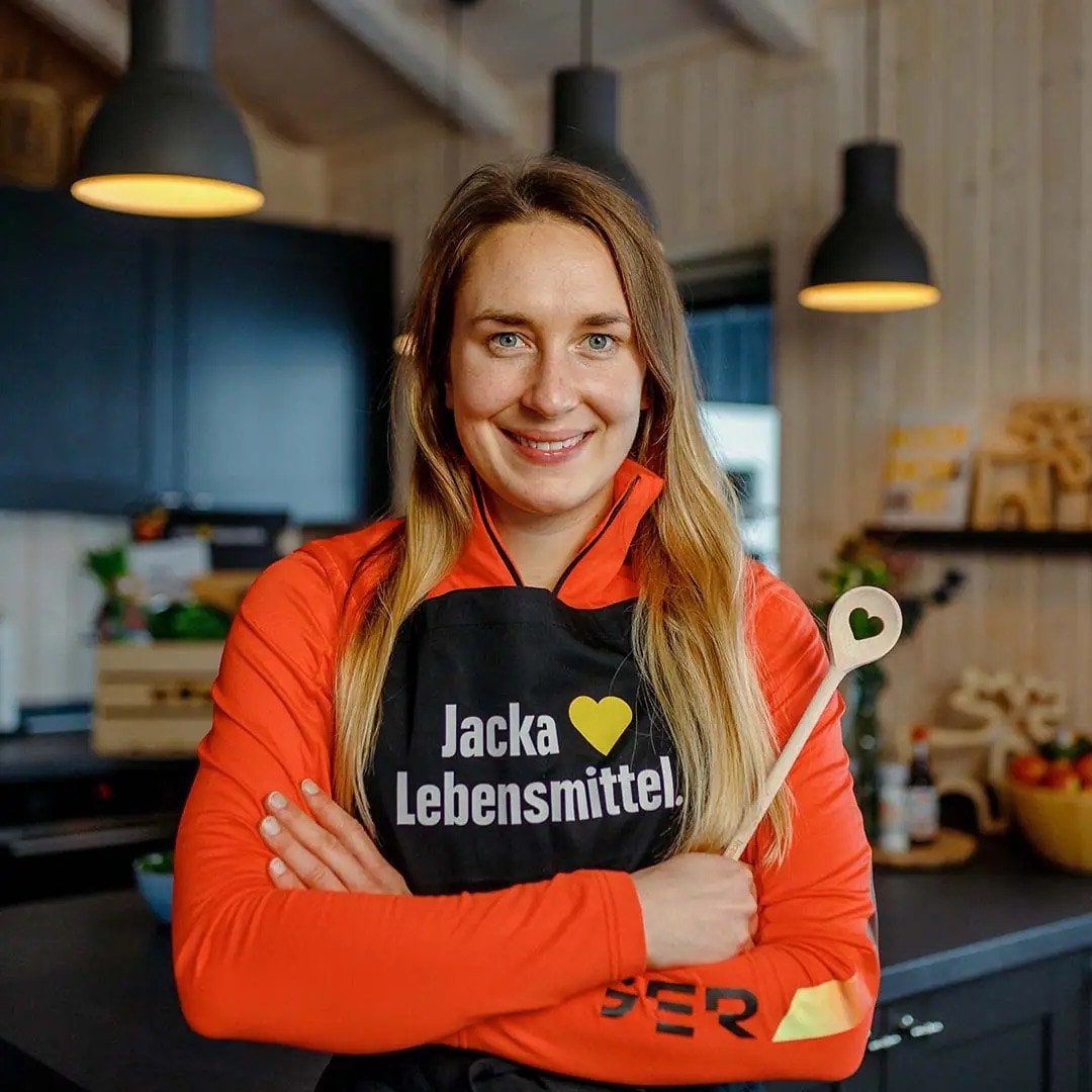 Junge Frau mit Sportjacke und schwarzer Schürze mit Aufschrift „Jacka liebt Lebensmittel.“ Sie hält einen hölzernen Kochlöffel vor sich.