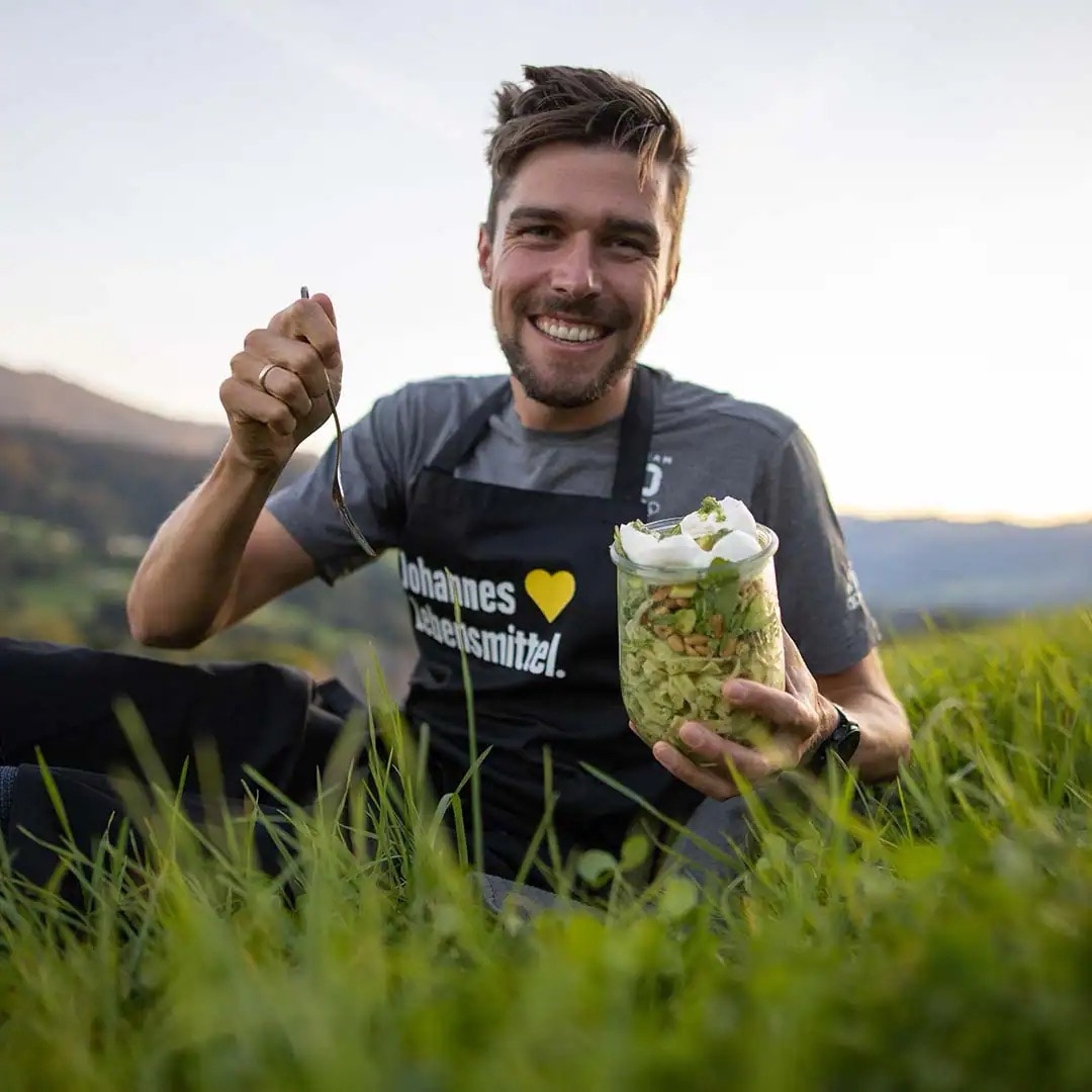 Junger Mann mit schwarzer EDEKA-Schürze steht auf einer grünen Wiese. Er hält eine Gabel und ein Einmachglas in den Händen.