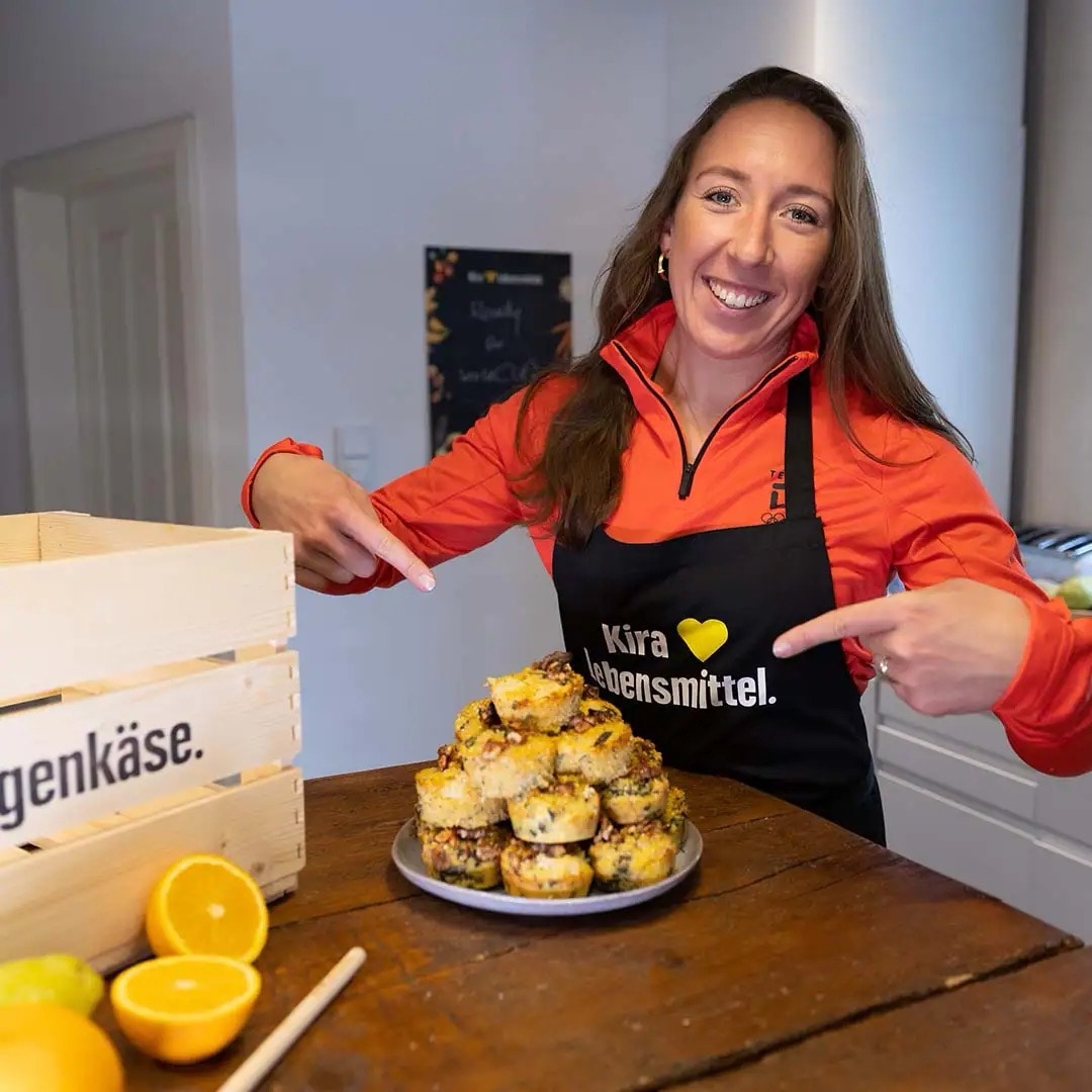 Eine junge Frau in roter Sportjacke und schwarzer EDEKA-Schürze, auf der „Kira steht Lebensmittel“ steht. Vor ihr auf dem Holztisch steht ein Teller mit Muffins.