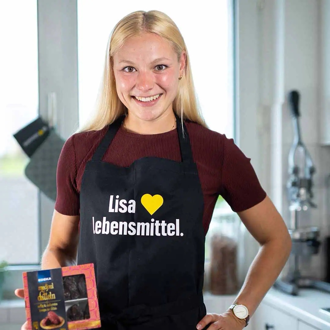 Junge Frau mit schwarzer EDEKA-Schürze, auf der „Lisa liebt Lebensmittel“ steht. In ihrer Hand hält sie eine Packung Datteln.
