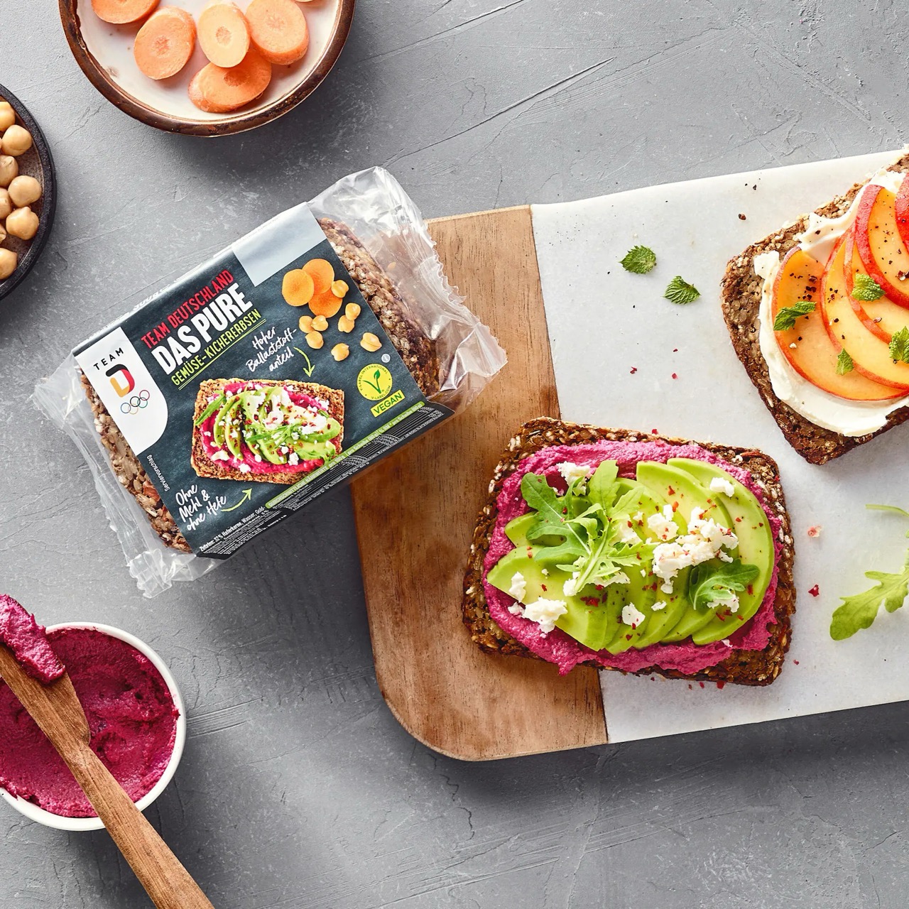 Eine Packung Schwarzbrot mit Gemüse und Kichererbsen. Daneben auf einem Brett zwei bunt belegte Scheiben des Brotes.