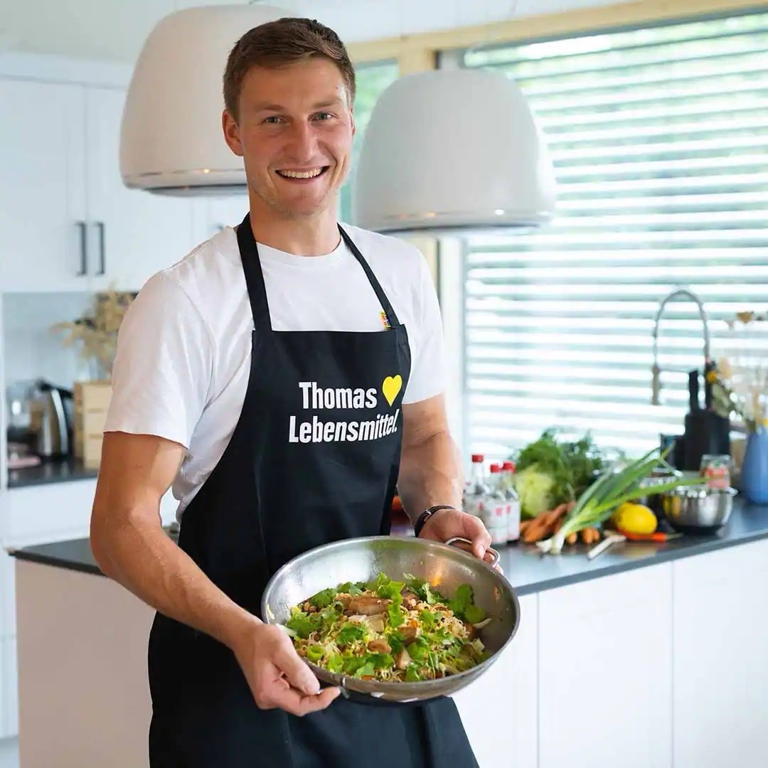 Junger Mann steht in einer Küche mit einer Salatschüssel aus Edelstahl in seinen Händen. Auf seiner schwarzen EDEKA-Schürze steht „Thomas liebt Lebensmitte.“