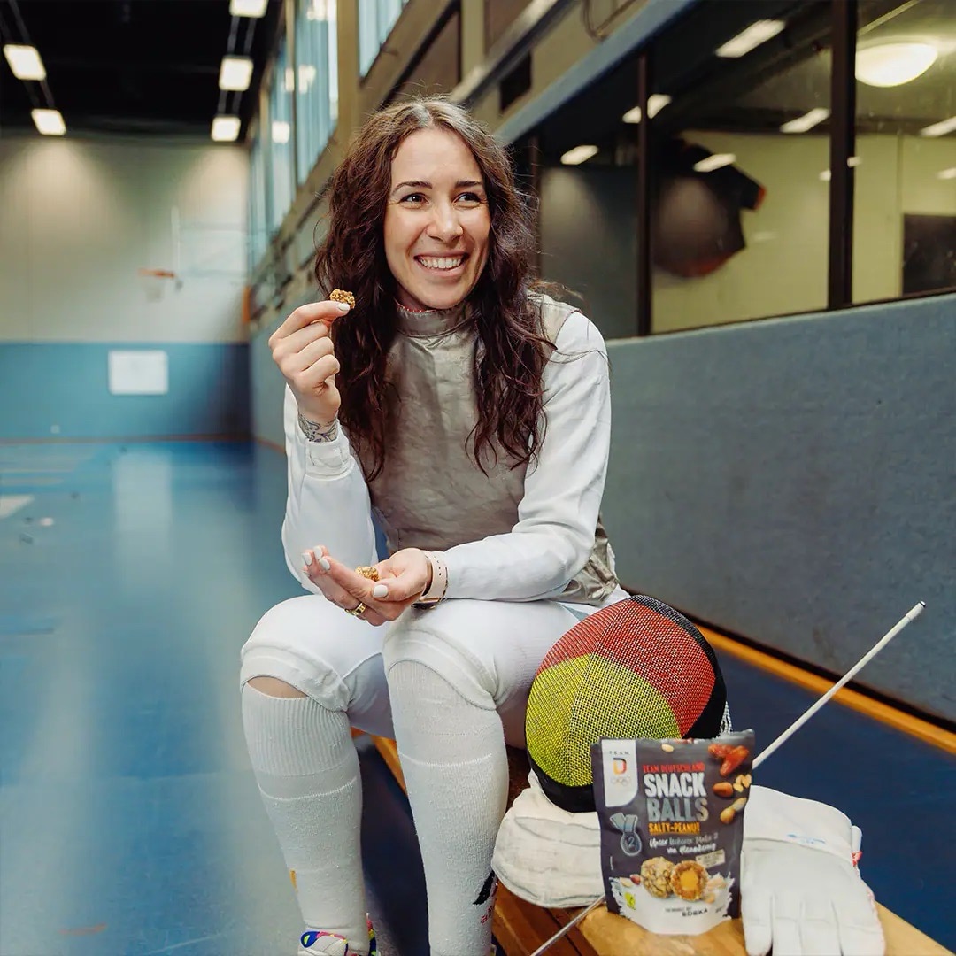 Junge Frau sitzt auf einer Bank in einer Sporthalle. Sie trägt weiße Fecht-Kleidung und hält einen Snack-Ball in der Hand.