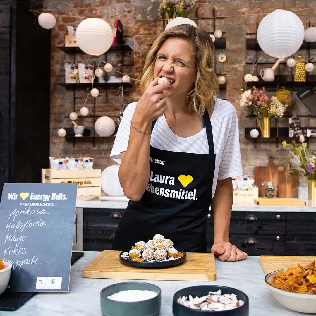 Junge Frau mit schwarzer Schürze beißt in einen Energy-Ball. Auf einer Tafel stehen die Zutaten: Aprikosen, Haferflocken, Mango, Kokos, Mangopaste.