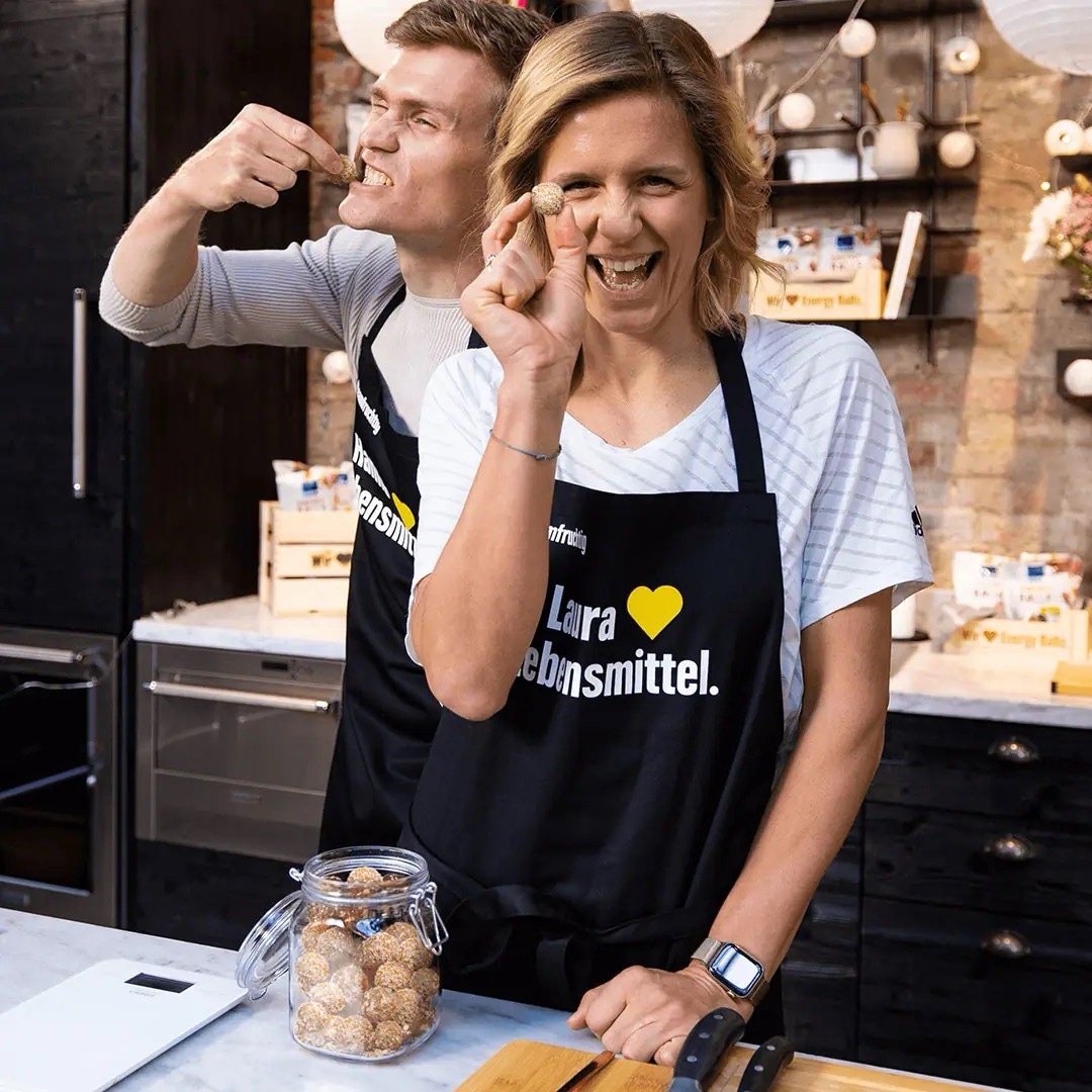 Lachende juge Frau hält einen Snack-Ball. Im Hintergrund beißt ein junger Mann in einen Snack-Ball.
