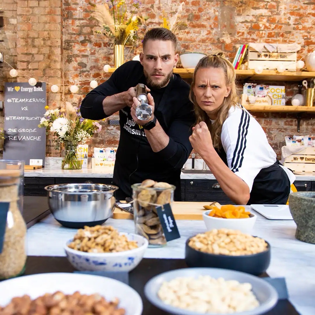 Ein Mann und eine Frau posieren vor einer Küchenzeile. Er hält einen Stabmixer, sie ballt die linke Faust.