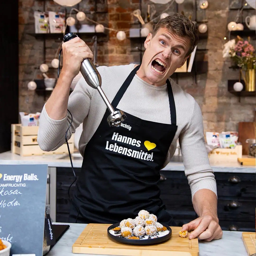 Junger Mann mit schwarzer EDEKA-Schürze. Er hält einen Stabmixer hoch über einem Teller Snack-Balls und verzehrt das Gesicht.