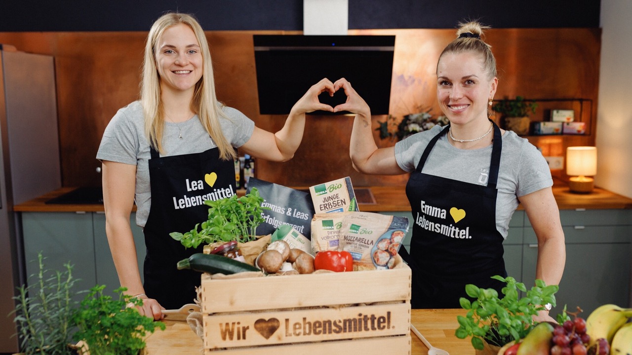 Zwei junge Frauen in EDEKA-Schürzen formen ein Herz mit jeweils einer Hand. Vor ihnen steht ein Holzkorb mit Lebensmitteln.
