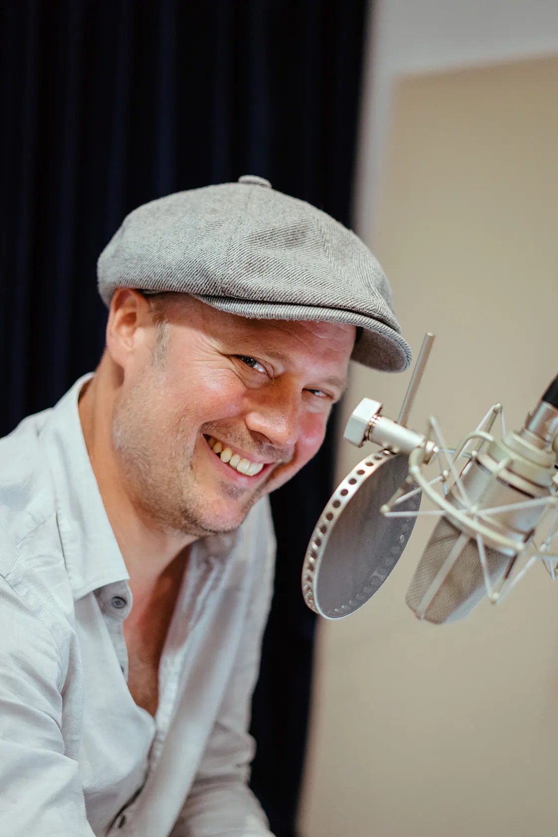 Ein lachender Mann mit heller Schiebermütze an einem Mikrofon mit Pop-Schutz.