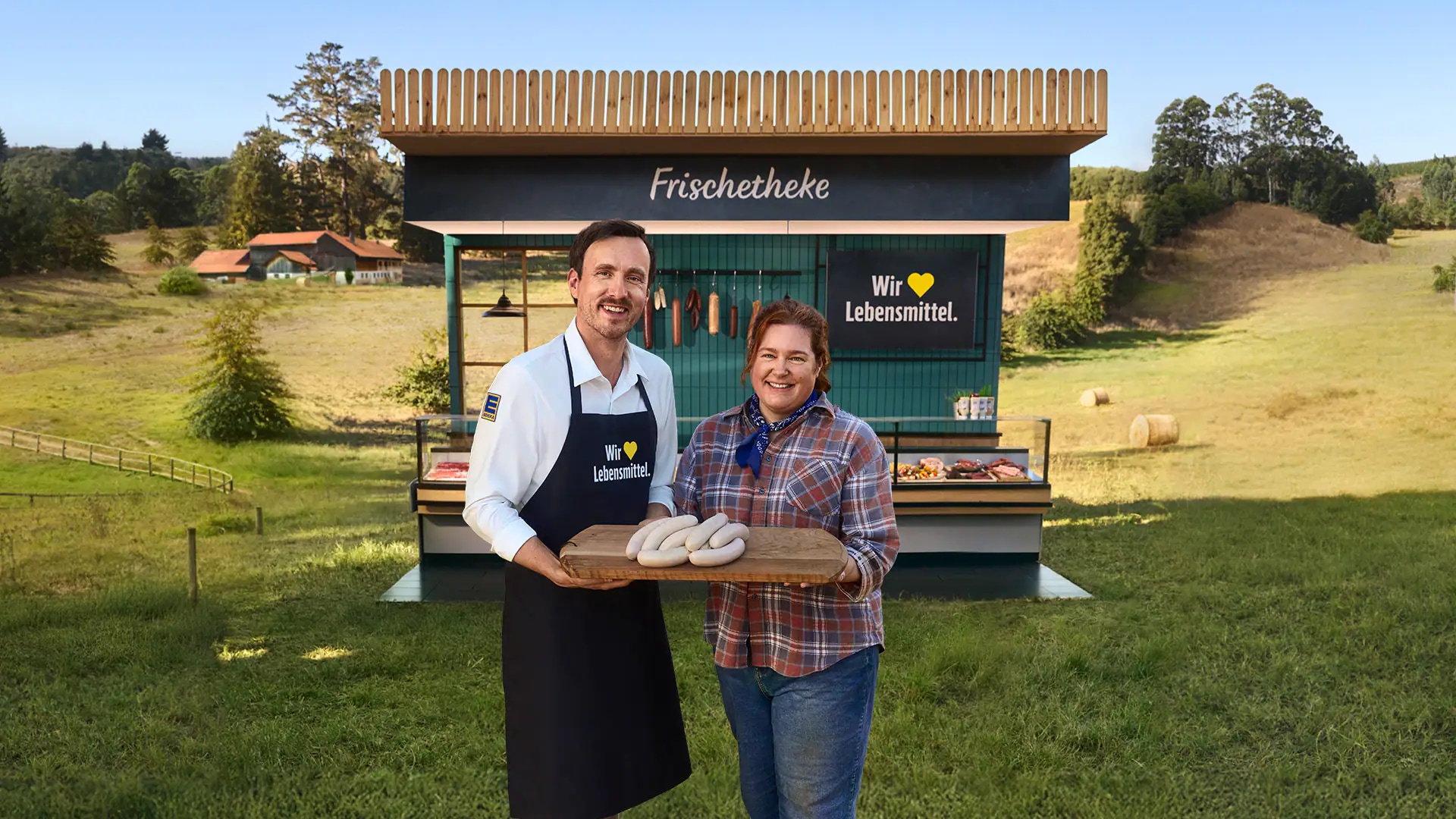 Ein Mann und eine Frau stehen vor einer EDEKA Frischetheke auf einer Wiese und halten ein Brett mit Würsten in den Händen. Der Mann trägt eine EDEKA Schürze.