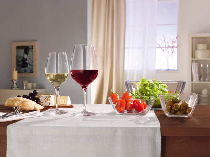 Gedeckter Tisch mit zwei Gläsern Rot- und Weißwein, dazu Salat, Kirschtomaten, Oliven, Käse und Brot in einer hellen Wohnumgebung.