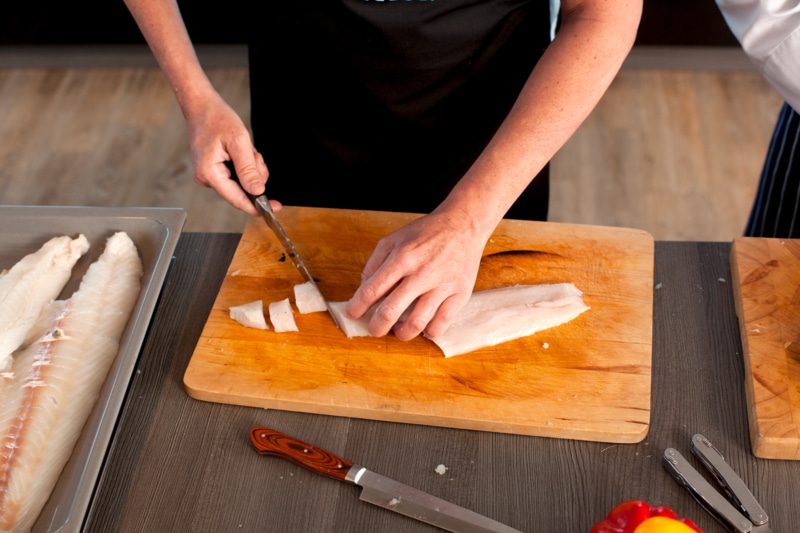 Fischfilet wird auf einem Schneidebrett mit einem Messer in Stücke geschnitten, daneben liegen weitere Filets und Küchenutensilien.