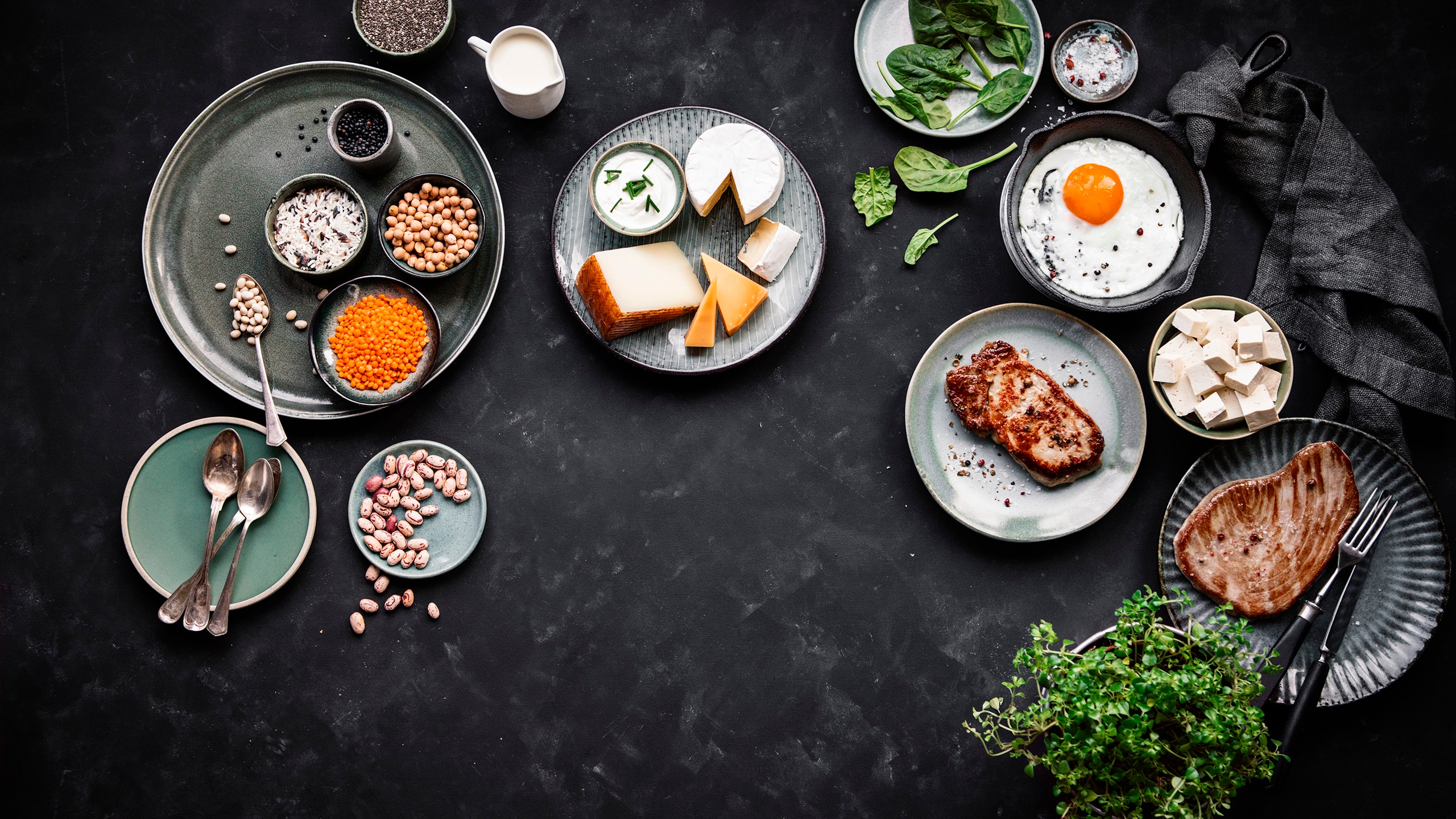 Verschiedene proteinreiche Lebensmittel wie Hülsenfrüchte, Käse, Ei, Tofu, Fleisch und Spinat auf einem dunklen Tisch arrangiert.