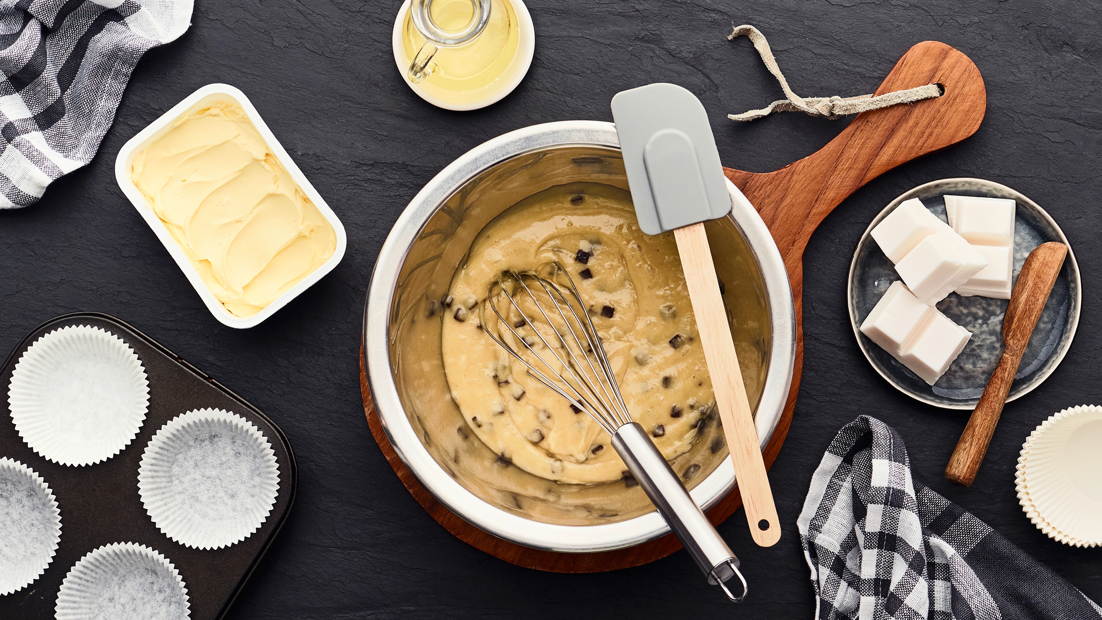 Schüssel mit Muffinteig und Schneebesen auf einer Küchenarbeitsfläche, umgeben von Backzutaten wie Butter, Zuckerwürfeln, Muffinförmchen und Teigschaber.