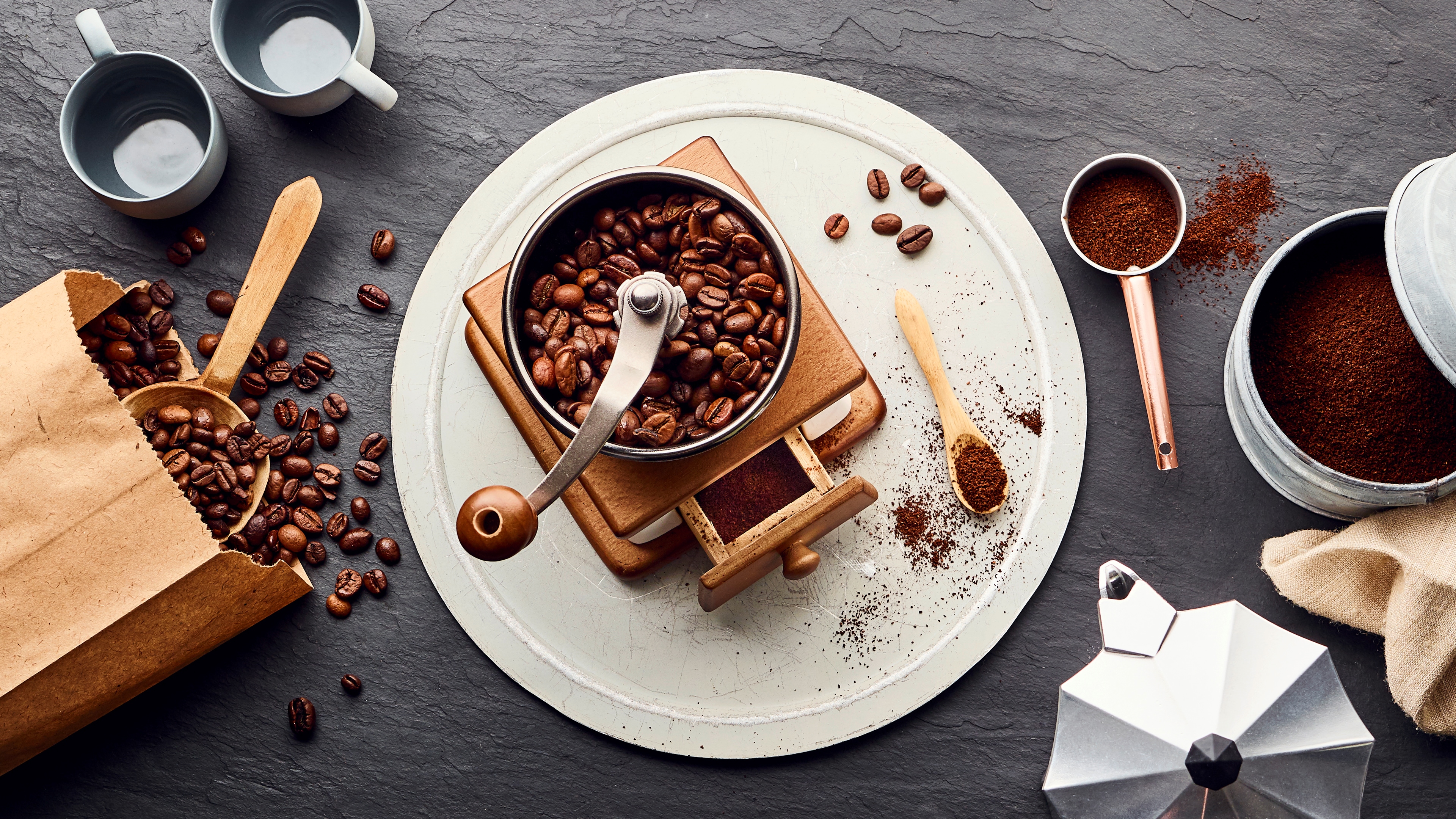 Manuelle Kaffeemühle auf weißem Teller, umgeben von Kaffeebohnen und gemahlenem Kaffee.