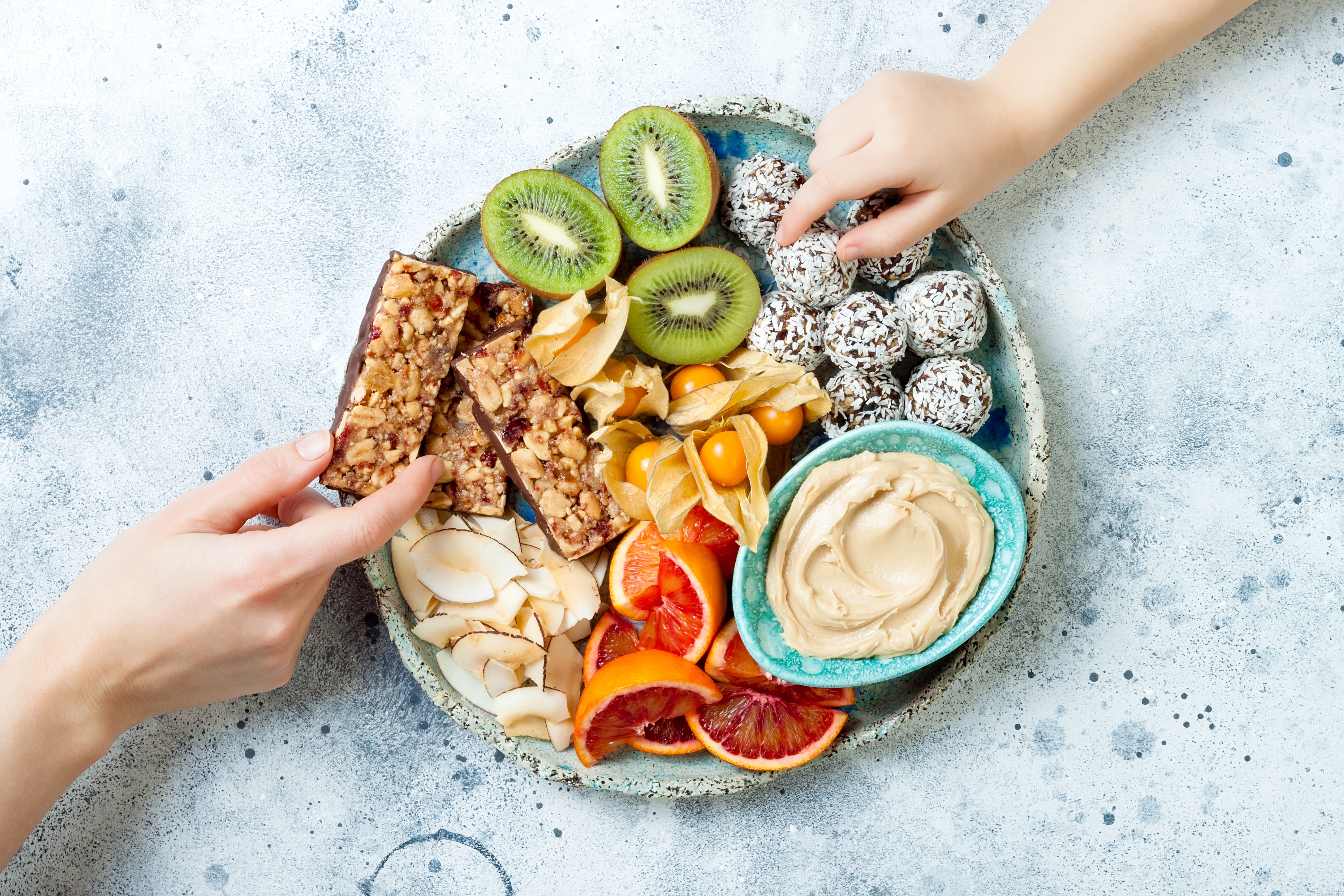 Teller mit veganen Lebensmitteln wie Müsliriegel, Hummus und Energiebälle mit einer Kinderhand und einer Erwachsenenhand, die danach greifen.