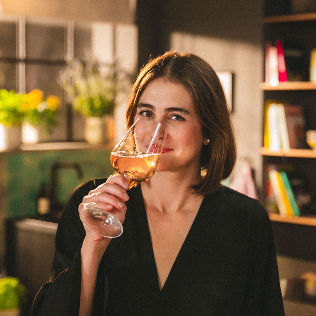 Person hält ein Glas Roséwein in einer Wohnküche; im Hintergrund sind Pflanzen, Regale und warme Beleuchtung zu sehen.