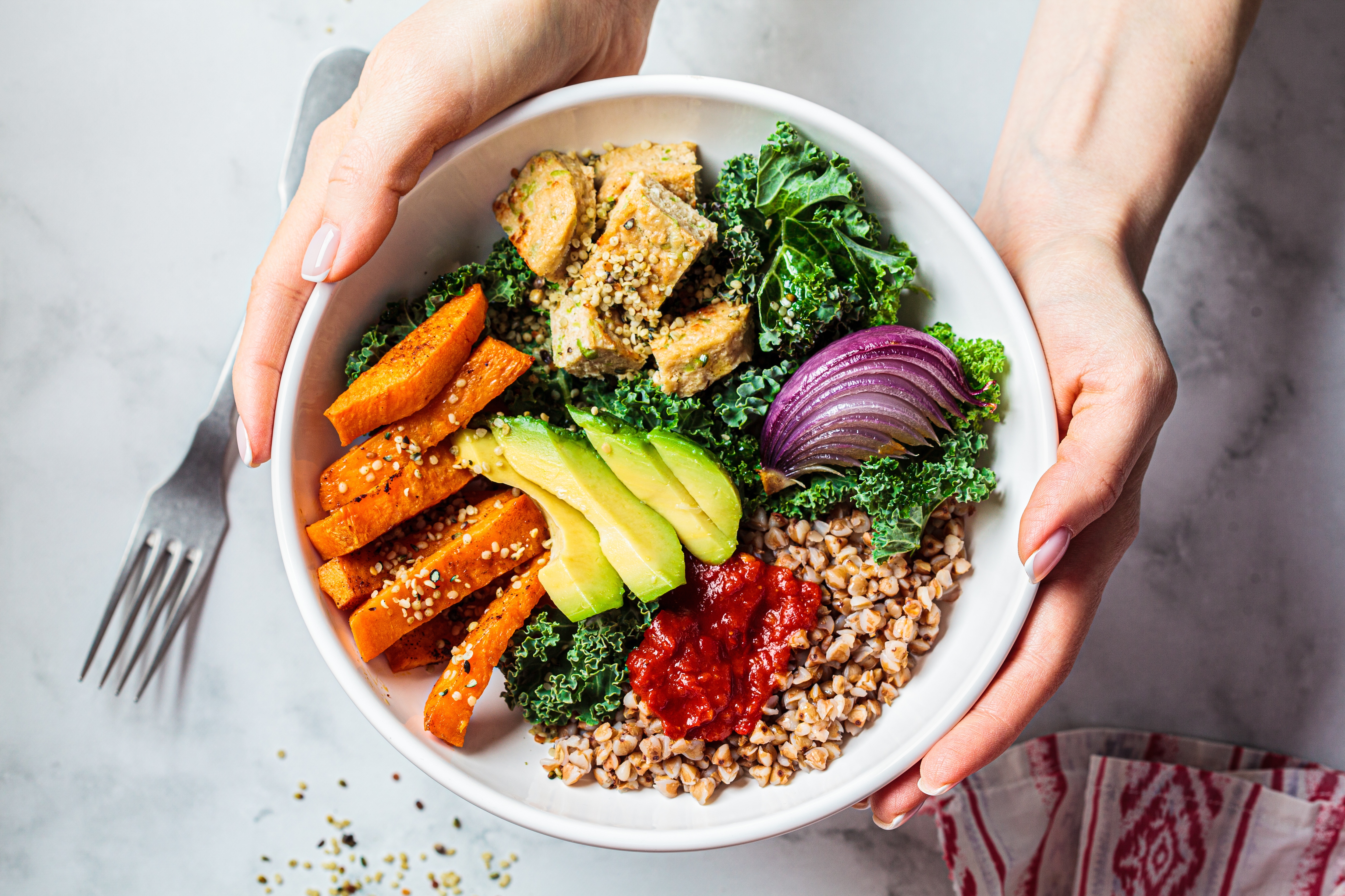 Hände einer Frau beim Essen eines veganen Salats – eine „Buddha Bowl“ mit gebackenem Gemüse, Avocado, Tofu und Buchweizen; Ansicht von oben. Konzept für pflanzliche Ernährung.