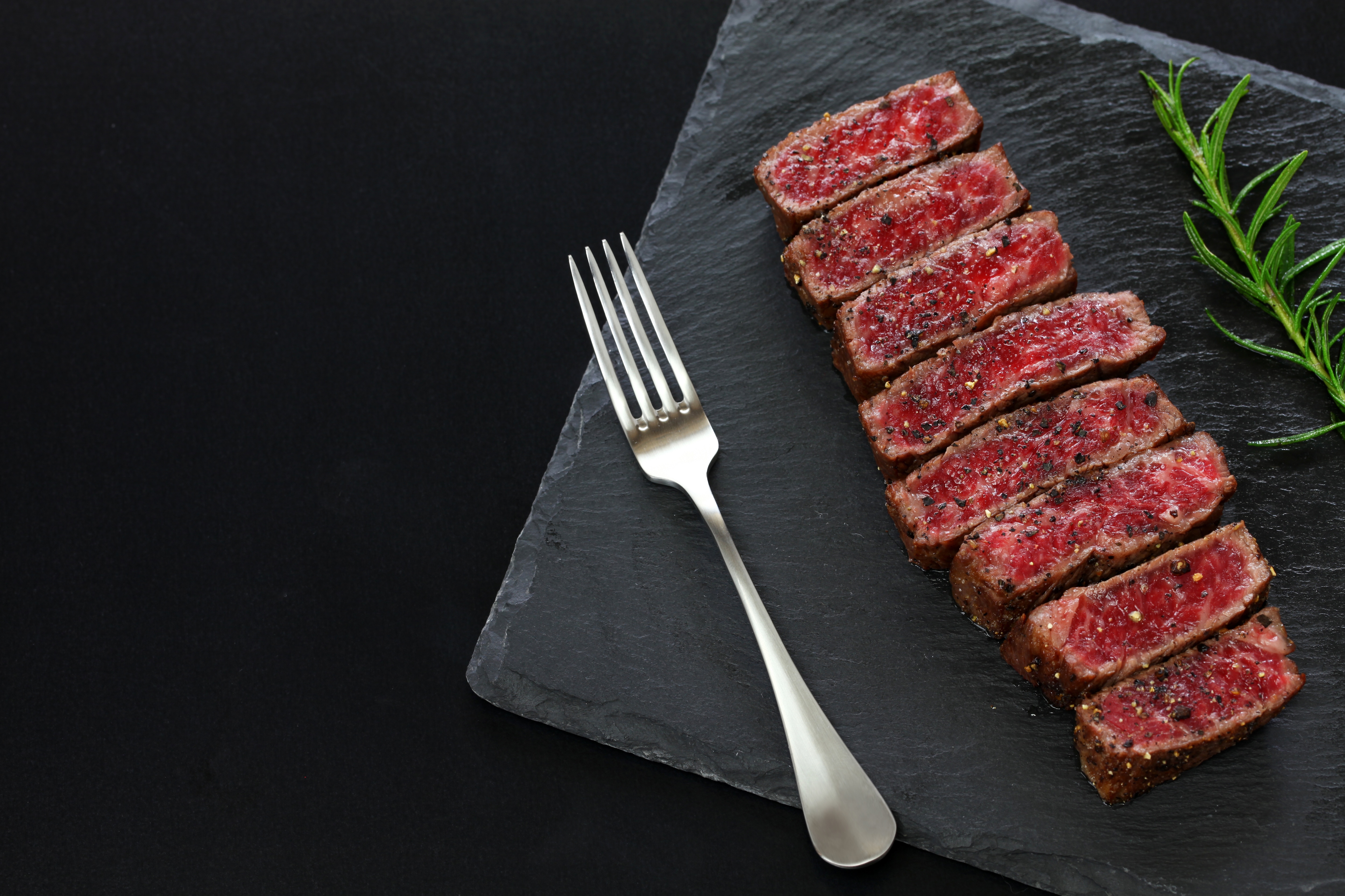 Aufgeschnittenes Steak mit rosa Kern auf schwarzer Schieferplatte, daneben eine Gabel und Rosmarin.